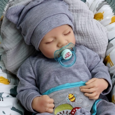 Bebé deitado com roupa cinzenta, chupeta azul com texto, cobertor cinzento bordado e lençóis estampados.