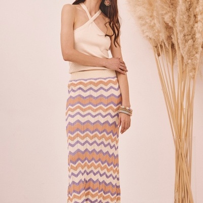 Mulher com top creme e saia comprida colorida em ambiente claro com plantas secas.