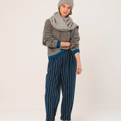 Mulher veste camisola cinzenta e cachecol grossos, calças largas azuis às riscas pretas, sapatilhas brancas e cinzentas e gorro cinzento