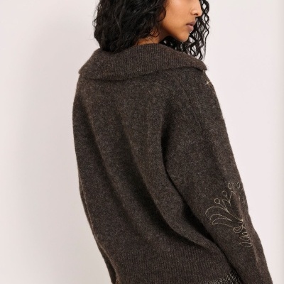 Camisola castanha de malha grossa com gola baixa e bordados dourados nas mangas, vestida por mulher de cabelos pretos encaracolados vista de costas.