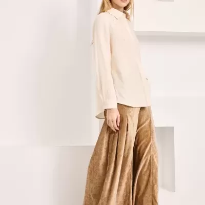 Mulher com camisa bege clara e calças largas castanhas num fundo branco.