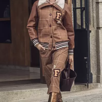 Mulher a usar casaco e calças castanhas às riscas com letra F, botas douradas, mala castanha e óculos de sol numa rua de pedra.