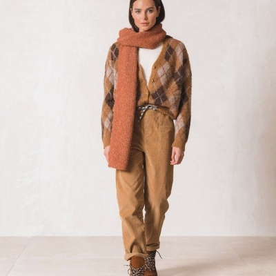 Mulher com cachecol castanho-alaranjado longo, camisola castanha com padrão de losangos, calças castanhas e botas castanhas