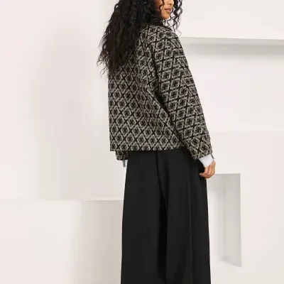 Mulher de costas com casaco geométrico castanho e preto, calças largas pretas e sapatos castanhos e pretos em ambiente interior branco.