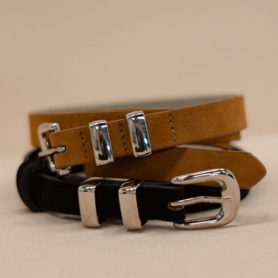 Dois cintos finos em pele castanho claro e preto com fivelas metálicas prateadas sobre superfície bege.