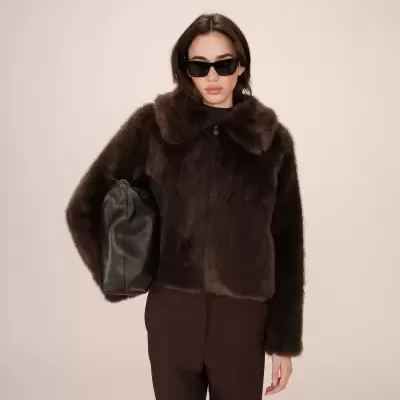 Mulher com casaco de pelo castanho, calças castanhas, óculos escuros pretos e bolsa de couro preto.