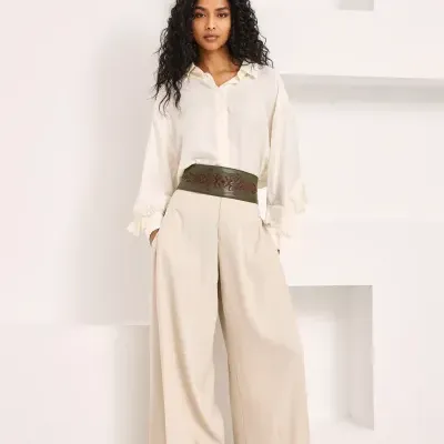 Mulher com camisa creme, calças largas bege, cinto verde decorado e botas pretas em fundo branco.