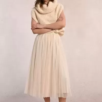 Mulher com colete de malha bege, saia plissada bege e ténis brancos com riscas rosa