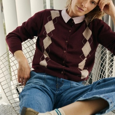 Mulher sentada numa cadeira metálica usando cardigan bordô de losangos branca e bege, camisa branca e jeans azuis.