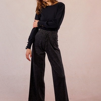 Mulher com camisola preta, calças largas pretas com brilho e sapatos castanhos sobre fundo neutro.