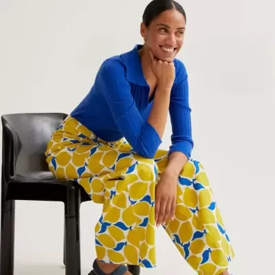 Mulher sentada com camisola azul, calças estampadas amarelas e azuis e sandálias azuis.