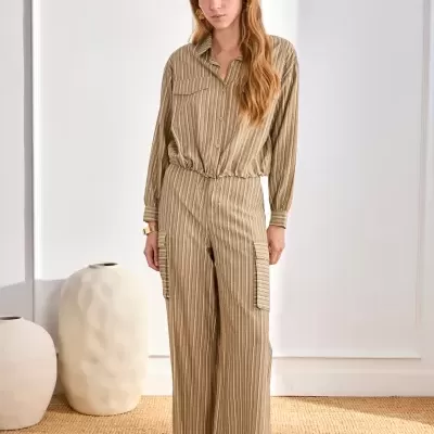 Mulher vestindo conjunto bege riscado com camisa e calças largas, sandálias castanhas, em ambiente claro com vasos grandes ao fundo.