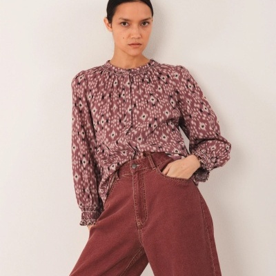 Mulher com blusa de padrão geométrico roxo e calças largas vinho em fundo branco