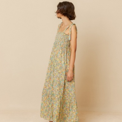 Mulher de vestido longo floral amarelo com sandálias castanhas em fundo bege
