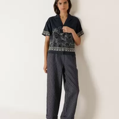 Mulher com camisa azul bordada e calças largas cinzentas em fundo branco