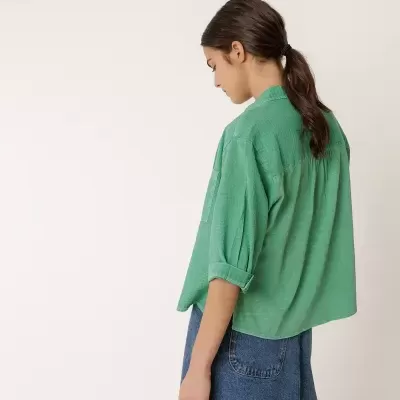 mulher de perfil e costas com camisa verde de manga 3/4 e saia azul de ganga