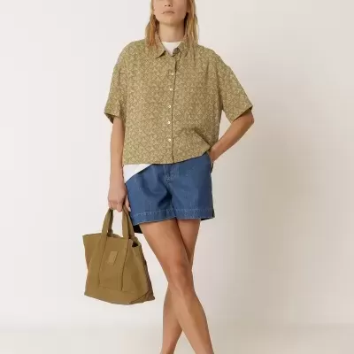 Mulher vestida com camisa bege estampada, calções de ganga azuis e a carregar uma bolsa de tecido mostarda