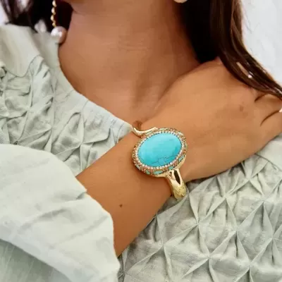 Pulseira dourada com pedra azul oval e cristais, vista no pulso de mulher com roupa verde clara.