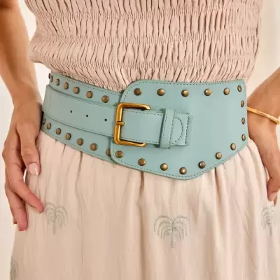 Cinto azul claro com tachas douradas e fivela dourada sobre vestido bege com bordado de palmeiras.