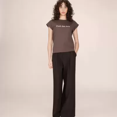 Mulher a usar camisola castanha com texto branco e calças pretas largas em fundo beje