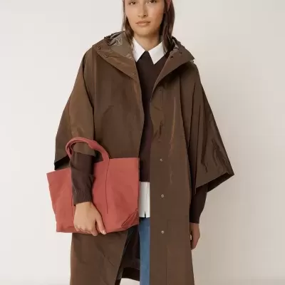 Mulher com capa impermeável castanha, malha castanha, camisa branca, jeans azuis e bolsa rosa de tecido