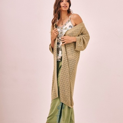 Mulher com cardigã bege trançado, blusa estampada branca, calças verdes e sandálias douradas