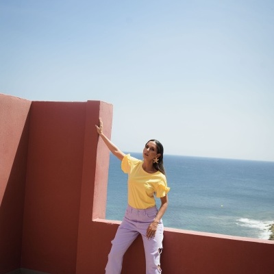 Mulher com calças lilás e camisola amarela num terraço com vista para o mar