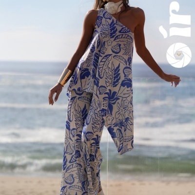 Modelo usa macacão azul e branco com estampa floral, sandálias brancas, e acessórios dourados junto ao mar.