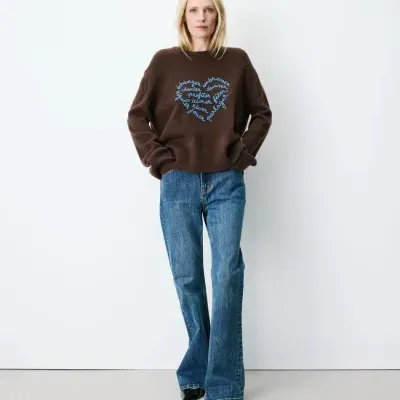 Mulher com camisola castanha de malha e calças jeans largas em fundo branco