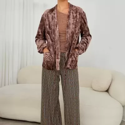 Mulher com casaco de veludo castanho, camisa castanha e calças largas com padrão geométrico, em interior com sofá creme.