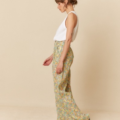 Modelo feminino com calças florais, top branco e sandálias pretas, de perfil num fundo bege.