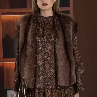 Mulher com vestido castanho de padrão cachemir, colete de pêlo castanho, óculos de sol quadrados e mala castanha.