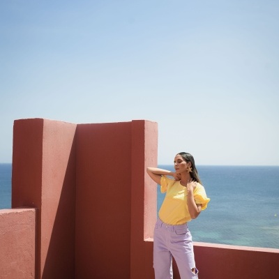 Mulher com t-shirt amarela e calças lilás junto a varanda vermelha com vista para o mar