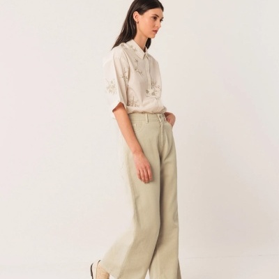 Mulher veste calças largas bege, camisa creme floral e sapatos brancos num fundo branco.