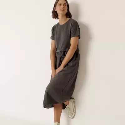 Mulher com vestido midi cinzento escuro e ténis brancos posando junto à parede