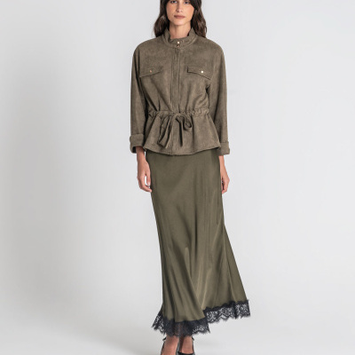 Mulher com casaco verde-oliva de camurça, saia longa verde com renda preta na barra e sapatos pretos de salto