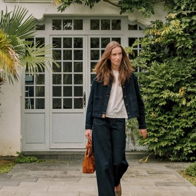 Mulher com casaco e calças de ganga azul escuro e mala castanha junto a porta de vidro