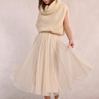 Mulher com colete de malha creme, saia plissada creme e ténis brancos com detalhes coloridos.