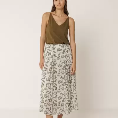 Mulher com top castanho, saia midi floral branca e sapatos cinza em fundo neutro.