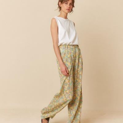 Mulher a usar calças florais largas, blusa branca sem mangas e sandálias pretas, em fundo bege