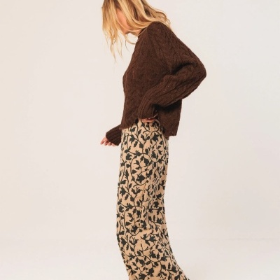 Mulher de perfil com camisola castanha de malha e calças largas bege com padrão de folhas pretas.