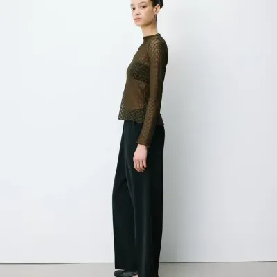 Mulher com blusa de renda castanha, calças largas pretas e sapatilhas negras em fundo branco.
