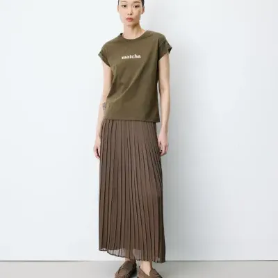 Mulher com t-shirt verde-oliva e saia plissada castanha em fundo branco.