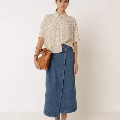 Mulher com blusa bege, saia jeans azul, sandálias castanhas e bolsa de couro castanha