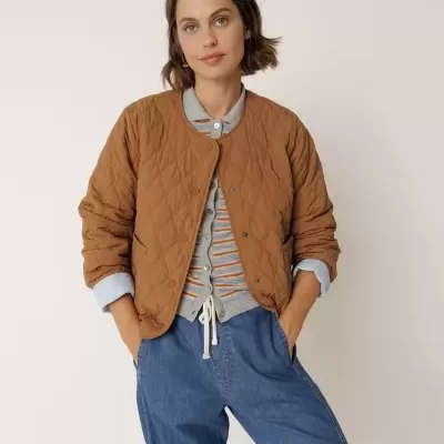 Mulher com casaco acolchoado castanho, camisa azul clara, camisola às riscas e calças de ganga azuis