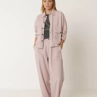 Mulher vestindo casaco e calças largas cor rosa claro com t-shirt preta e sapatos cinzentos em fundo branco