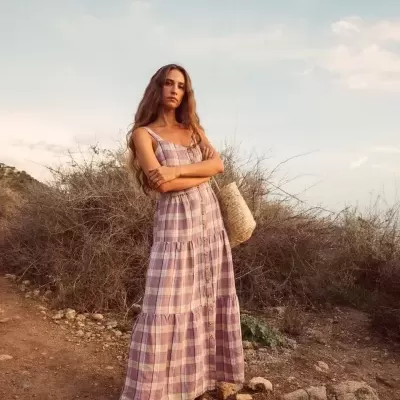 Mulher com vestido longo às quadrículas lilás e rosa, sandálias castanhas e bolsa de palha em cenário natural seco