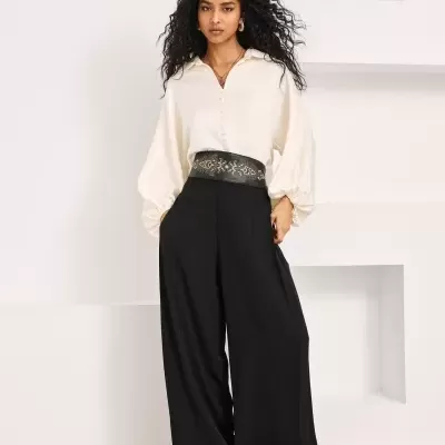 Mulher com calças pretas largas, blusa branca e botins dourados em ambiente interior minimalista