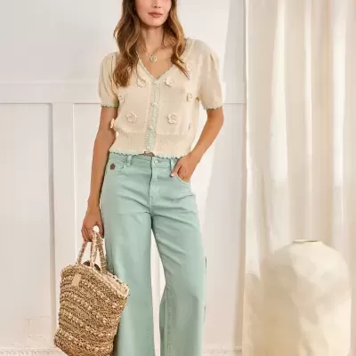 Mulher com camisola creme de malha com flores 3D e calças largas azul claro, com mala de palha e sandálias