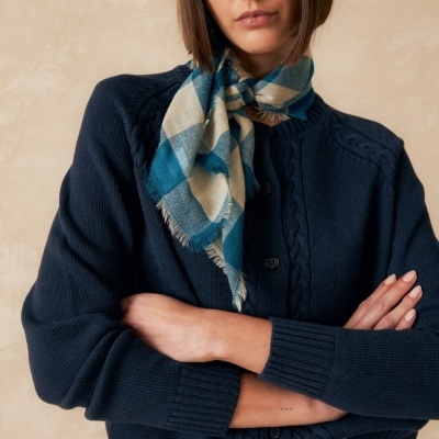 Mulher com cardigan azul escuro, lenço xadrez azul e bege, boné preto e calças de ganga